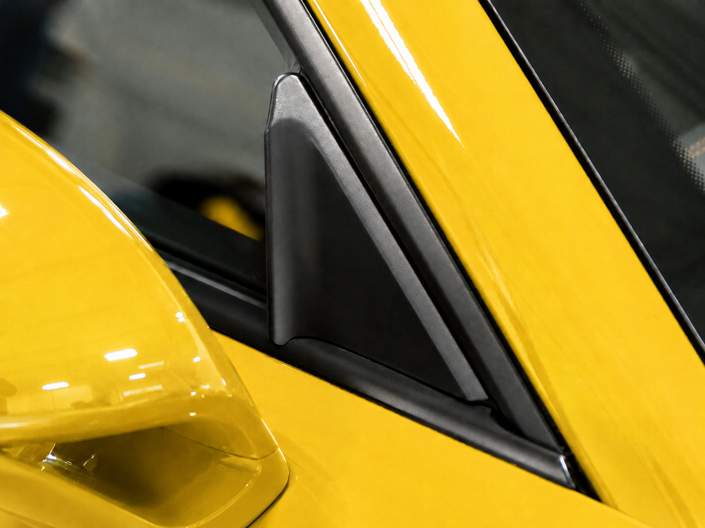 Black side window wind buffeting deflector detail fitted on the mirror trim area of a yellow Porsche 991